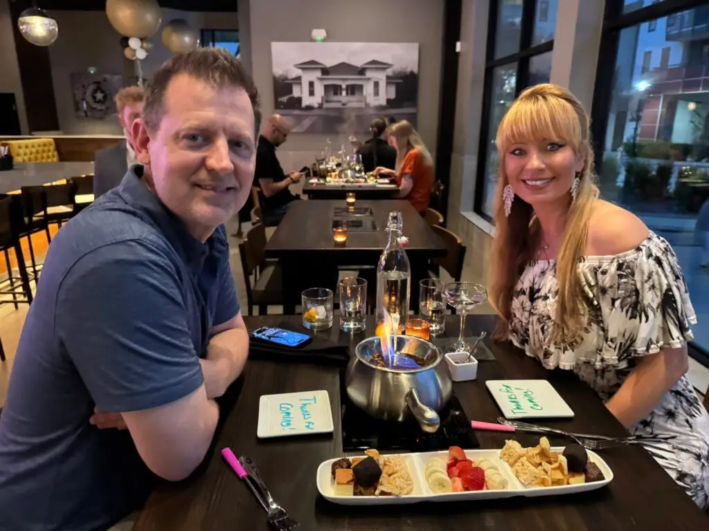 Smiling couple sits across from each other at a table with a flaming fondue pot, fruit and dessert dippers at The Melting Pot in Allen, Texas.