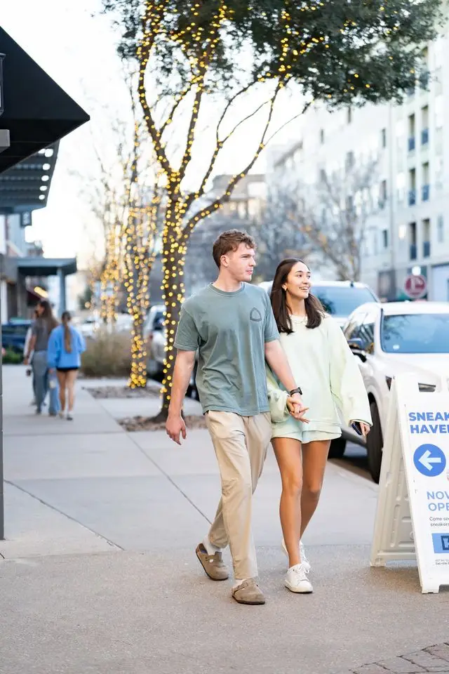 Couple holds hands while walking together along a tree-lined sidewalk at Watters Creek Village in Allen, Texas.