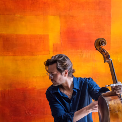 Cellist Joseph Kuipers holding his cello against a vibrant orange and yellow abstract background.