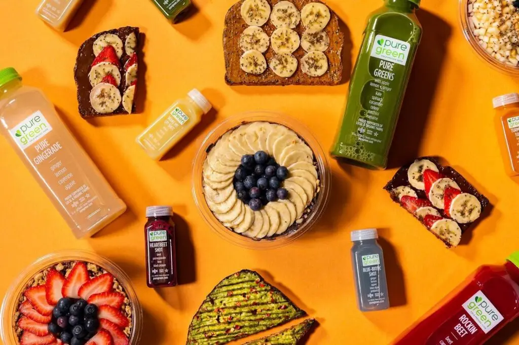 Overhead display of Pure Green cold-pressed juices, wellness shots, fruit-topped bowls, avocado toast, and toast topped with bananas and strawberries on a bright orange background.