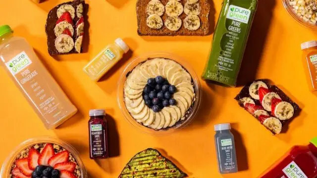 Overhead display of Pure Green cold-pressed juices, wellness shots, fruit-topped bowls, avocado toast, and toast topped with bananas and strawberries on a bright orange background.
