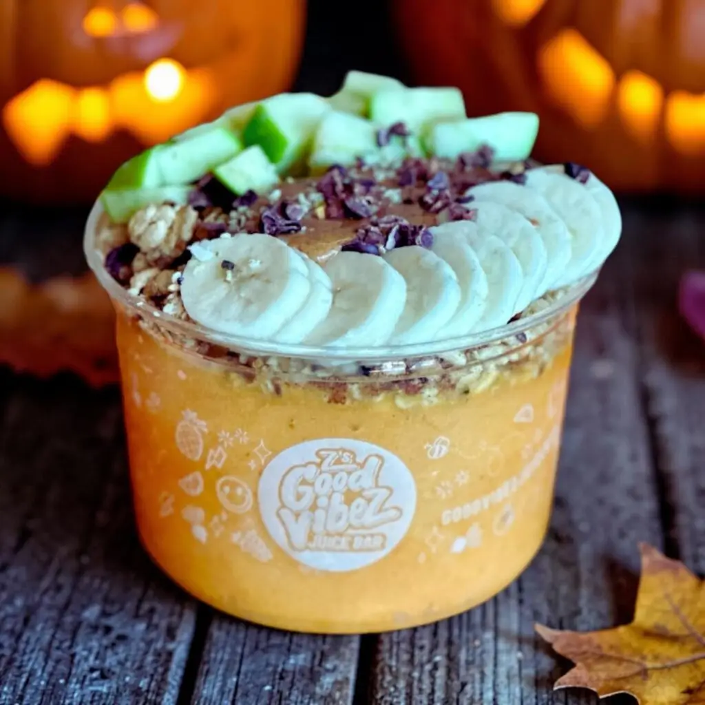 Pumpkin Patch Bowl from Z’s Good Vibez Juice Bar topped with banana, green apple, granola, almond butter and cacao nibs, with glowing jack-o’-lanterns in the background.