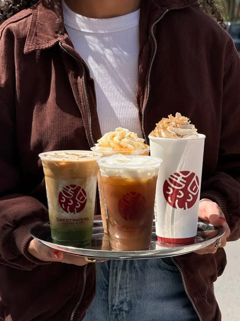Person holding a tray of four seasonal Sweetwaters Coffee & Tea drinks, including iced and whipped-cream-topped beverages.