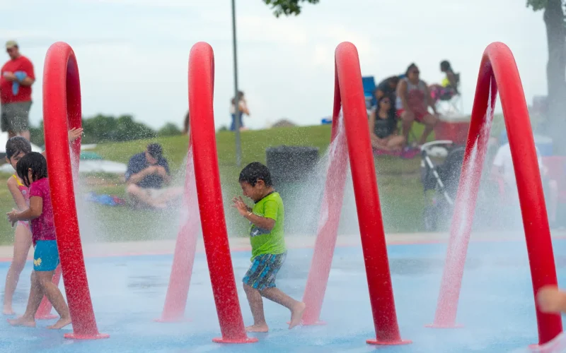 Children running through water sprays at a colorful outdoor splash pad with red arches in a public park.