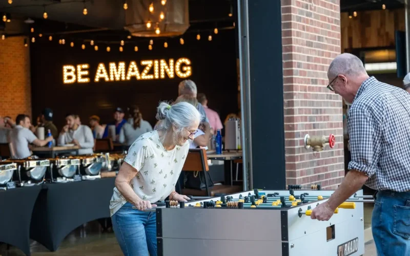 Couple plays foosball together at Chicken N Pickle in Allen, Texas, with other guests and a glowing “Be Amazing” sign in the background.