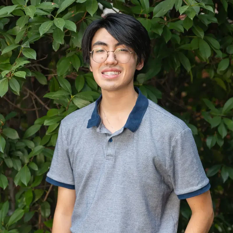 Portrait of Carlos smiling outdoors in front of green foliage, wearing glasses, a gray polo shirt and khaki pants.