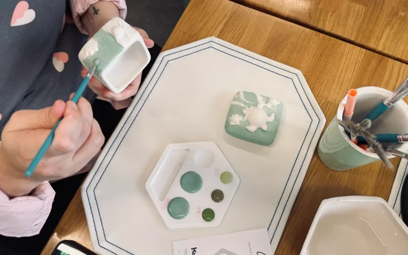 Close-up of hands painting a ceramic mug during a pottery painting session at Color Me Mine in Allen, Texas, with paint brushes, glaze colors, and a ceramic plate on a wooden table.
