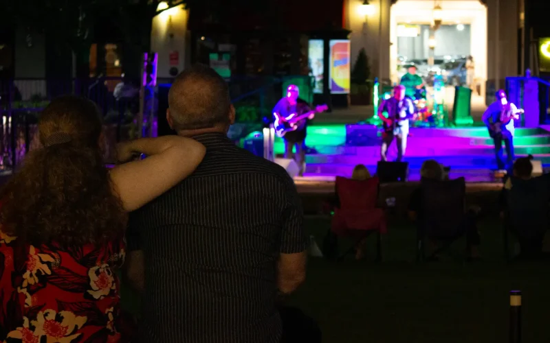 Guests enjoy live music during Concerts by the Creek at Watters Creek Village in Allen, Texas, as a couple watches a band perform on the Village Green stage while the audience relaxes on lawn chairs.