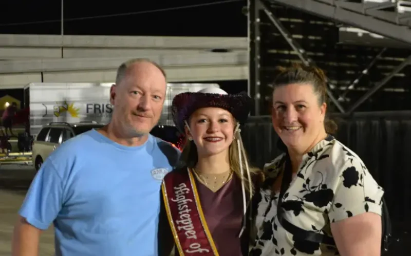 Teen wearing sash and cowboy hat poses with two adults at a community event at night