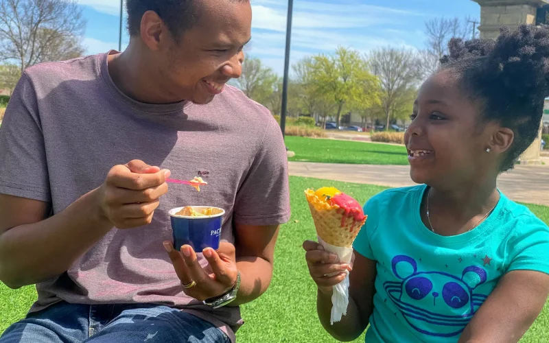 An adult and child enjoy ice cream together at The Village at Allen in Allen, Texas, smiling as they share a treat outdoors on a sunny day.