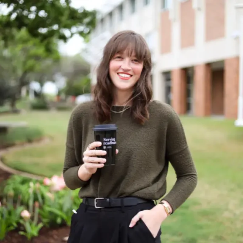 Rachel Alexander, Strategic Projects Manager, smiling outdoors while holding a coffee cup in front of a landscaped building setting