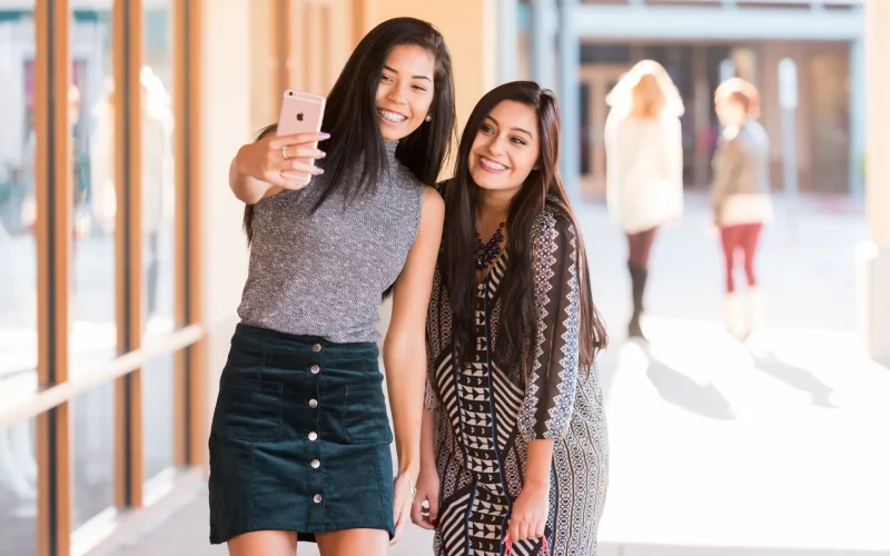 Two friends take a selfie while shopping at Allen Premium Outlets in Allen, Texas, with storefronts and other shoppers visible in the background.